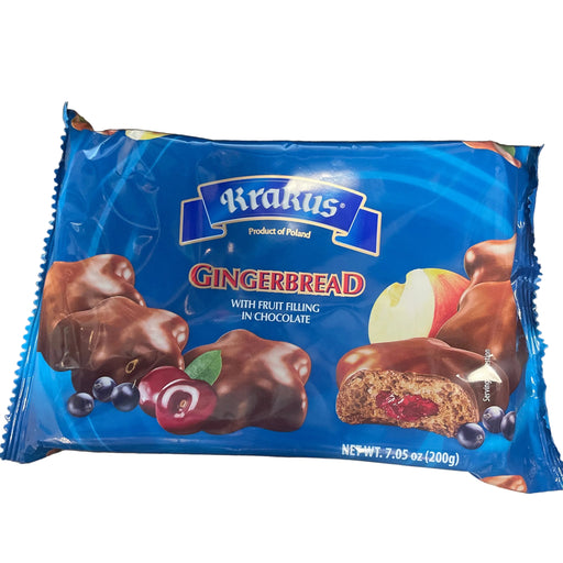 A package of Kirkus Gingerbread cookies with fruit bling in chocolate, placed on a dark wooden tabletop. The packaging is bright blue with illustrations of chocolate-covered gingerbread cookies, cherries, blueberries, and an apple slice.
